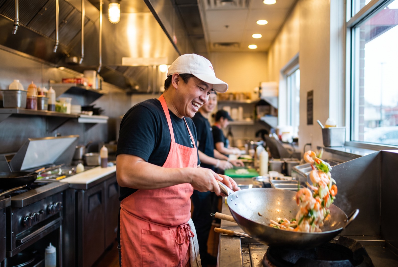 Line cook in a coral apron tossing shrimp in a wok