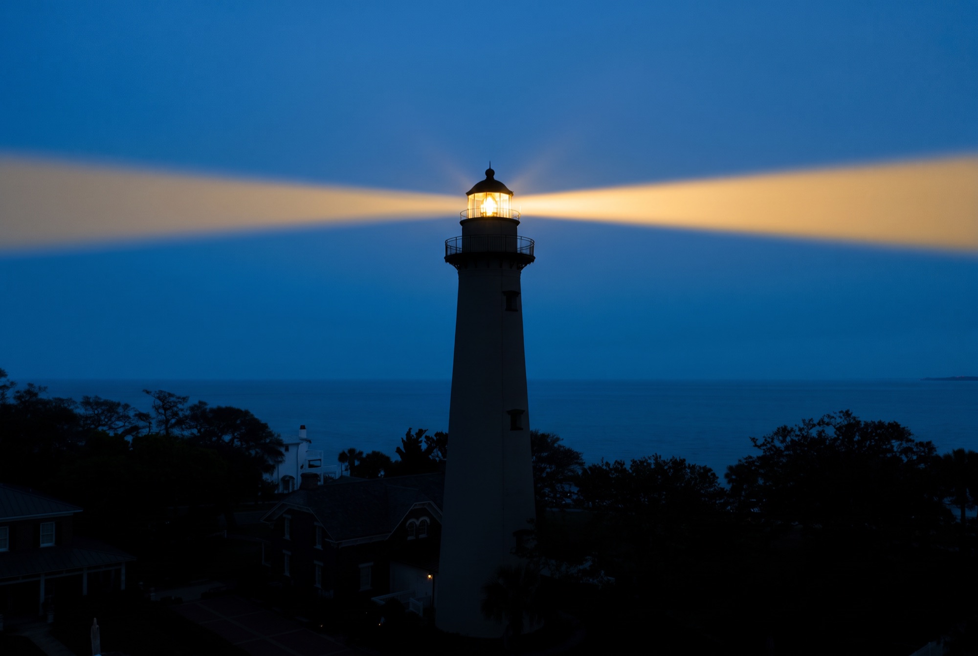 The St. Simons lighthouse at dusk, its warm gold beam cutting across a deep blue sky