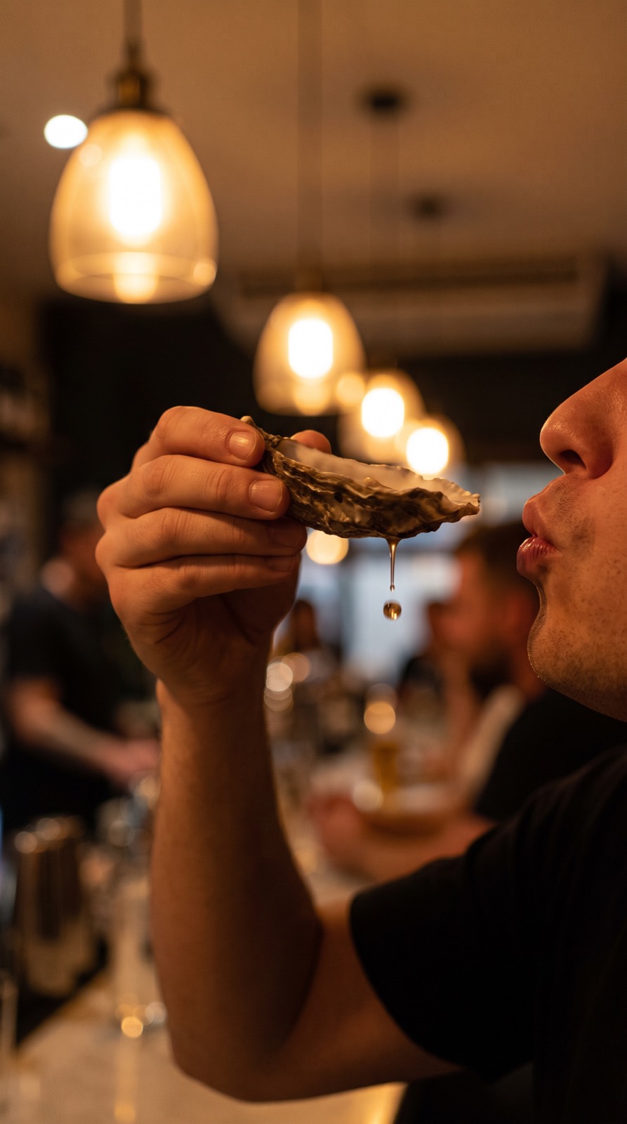Hand tipping an oyster shell toward the lips, bar bokeh behind