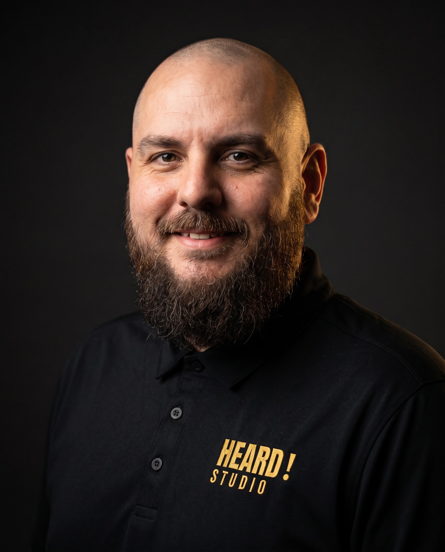 Portrait of Tyson Smith in dramatic low-key light, wearing a black polo with the gold Heard! Studio mark stitched on the chest
